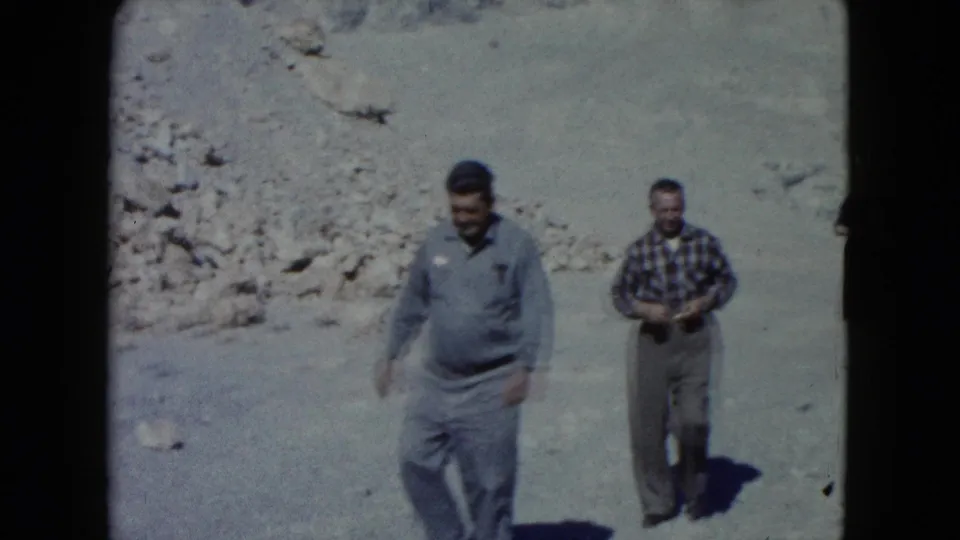 Two adult men, one in a blue shirt and one in plaid, walk through rocky Nevada desert terrain in 1960s 8mm footage.