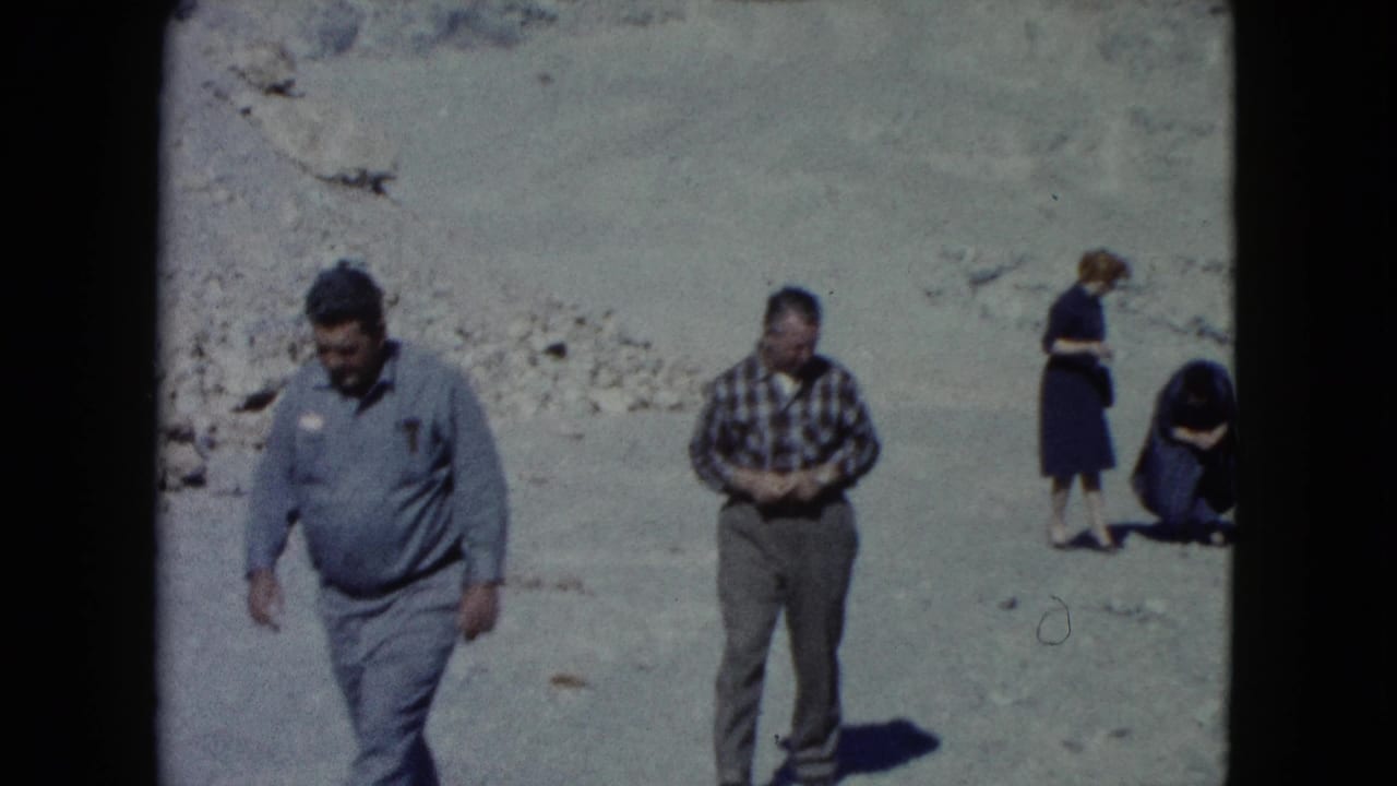 In Nevada 1960 home-movie footage, adults move across a barren desert wash with pale slopes behind them.