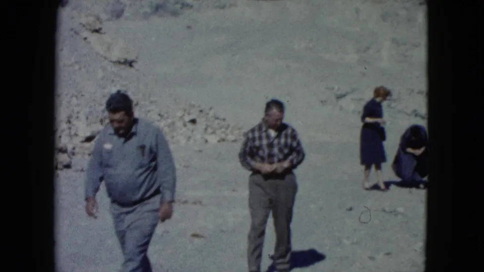 In Nevada 1960 home-movie footage, adults move across a barren desert wash with pale slopes behind them.