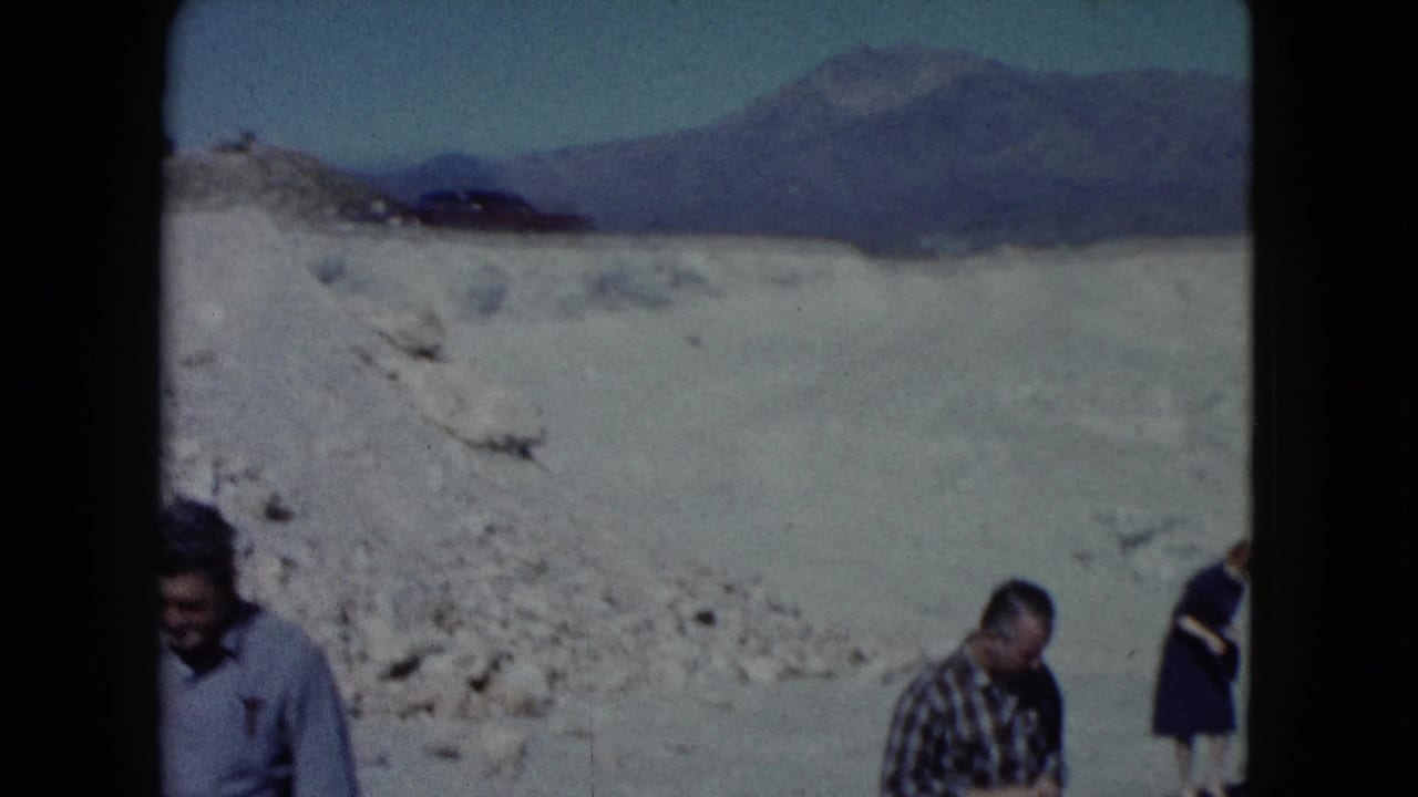 Adults appear near the edges of a wide desert view with pale ground, distant mountains, and blue sky.