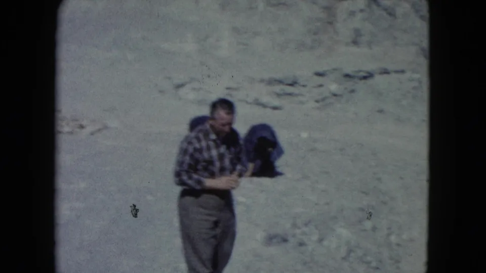 A man in plaid 1960s casual clothing pauses in the Nevada desert, with pale rocky ground filling the frame.