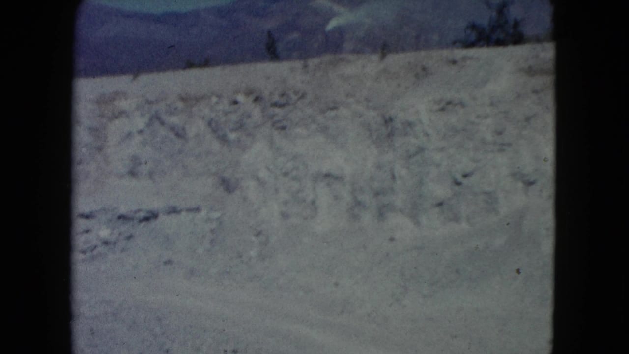 1960 Nevada 8mm frame showing a dry dirt slope, scattered scrub, distant mountain ridge, and blue sky.
