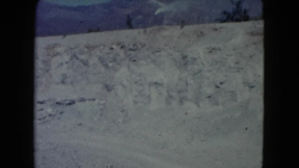 1960 Nevada 8mm frame showing a dry dirt slope, scattered scrub, distant mountain ridge, and blue sky.