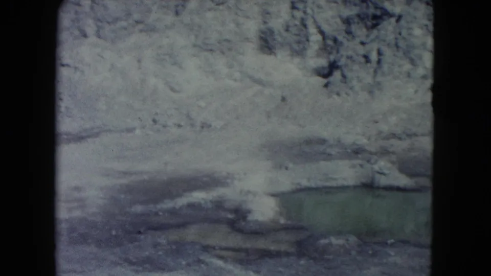 1960 Nevada 8mm frame showing pale rocky terrain, snow or ice patches, and a greenish pool near the lower edge.