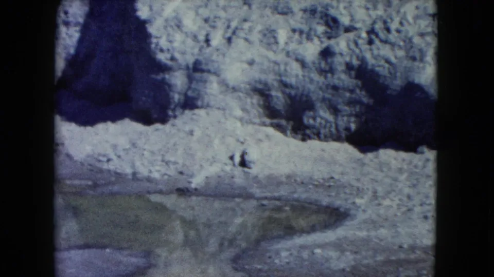 1960 Nevada 8mm frame showing a shadowed cave mouth, rough snow-dusted rock, and greenish water along the lower edge.