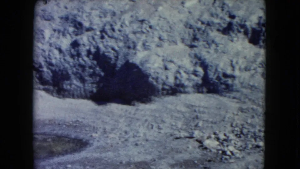 1960 Nevada 8mm frame showing rough winter rock, a dark cave-like opening, and a small greenish pool at the lower left.