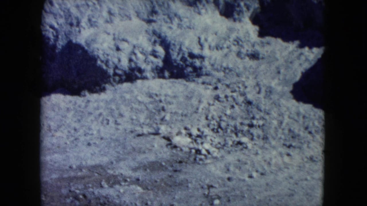 1960 Nevada 8mm frame showing pale rocky desert terrain, scattered stones, and dark shadowed erosion under soft focus.