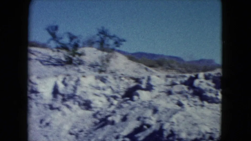 1960 Nevada 8mm frame showing rocky desert ground, a small shrub, distant hills, and clear blue sky.