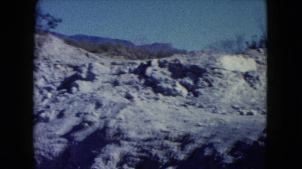 1960 Nevada 8mm frame showing pale rocky desert ground, sparse brush, and distant mountain ridges beneath a clear blue sky.