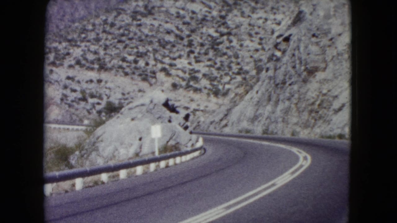 1960 Nevada road scene with a paved two-lane highway bending around pale rock cuts and scrub-covered desert slopes.