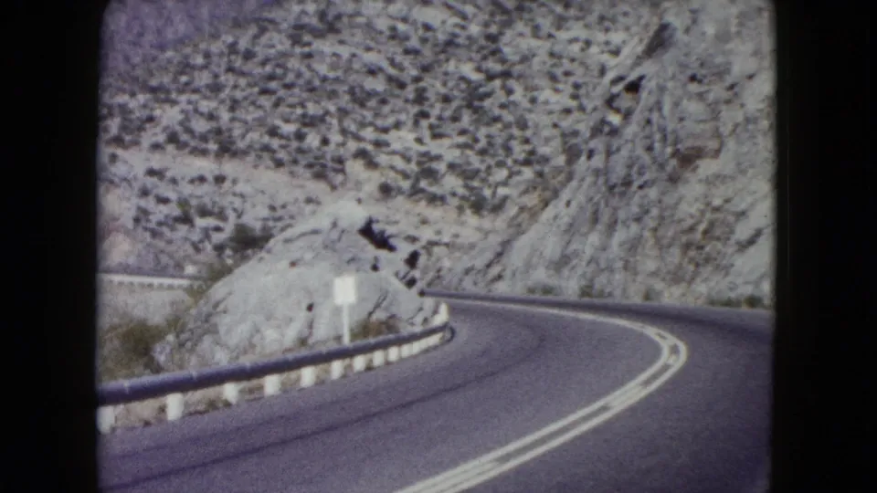 1960 Nevada road scene with a paved two-lane highway bending around pale rock cuts and scrub-covered desert slopes.