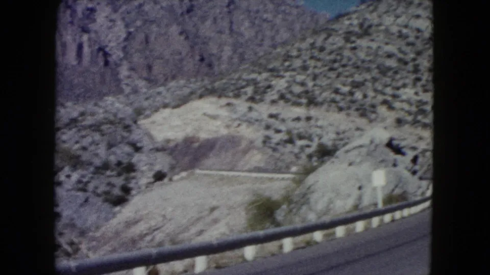 Forward-moving 8mm view of a Nevada mountain road edge, with guardrail, road surface, pale rocks, and sparse desert vegetation.