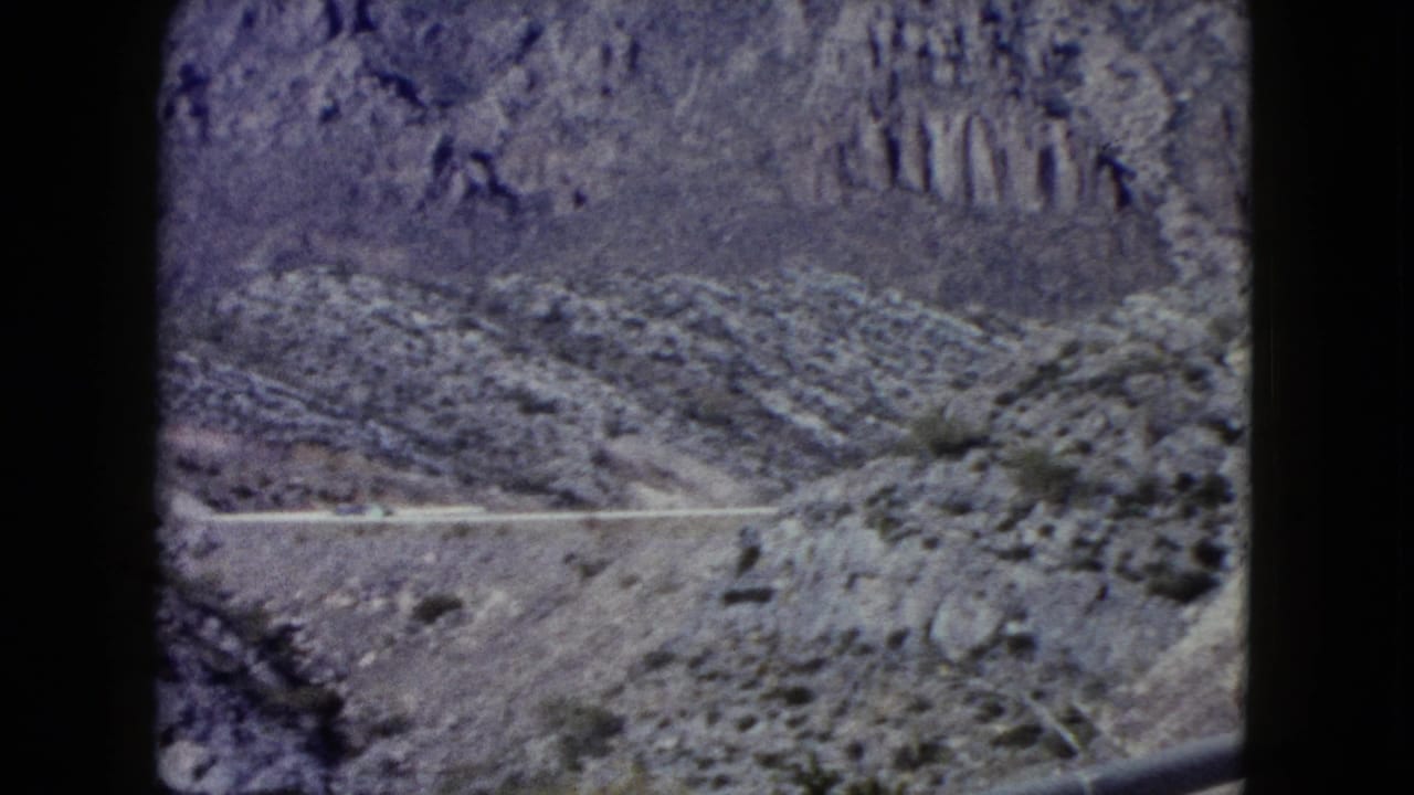1960 Nevada 8mm frame showing a distant roadway cutting across rocky desert hills beneath a steep mountain slope.