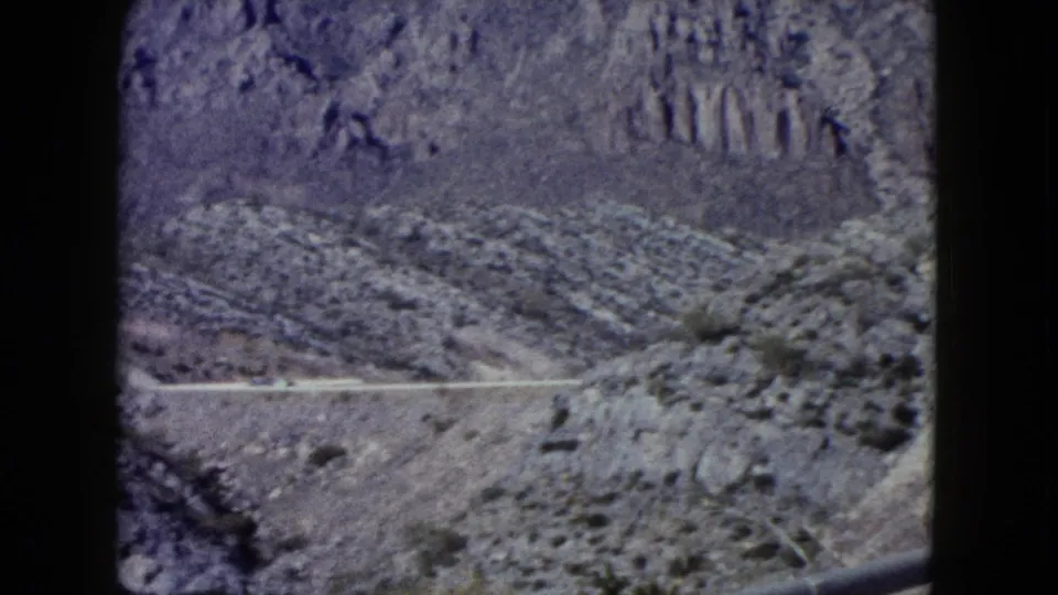 1960 Nevada 8mm frame showing a distant roadway cutting across rocky desert hills beneath a steep mountain slope.