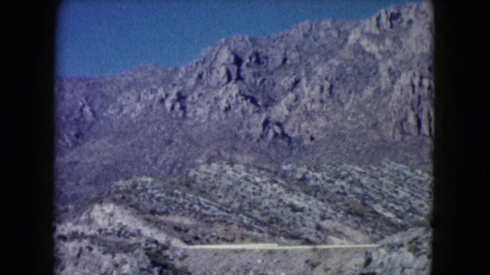 1960 Nevada 8mm frame showing scrubby hills, rocky ridges, and a pale road line across the landscape.
