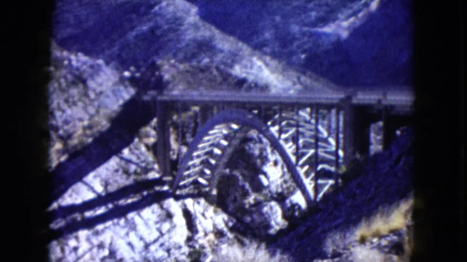 1960 Nevada 8mm frame showing a steel arch bridge set against steep rocky canyon terrain.