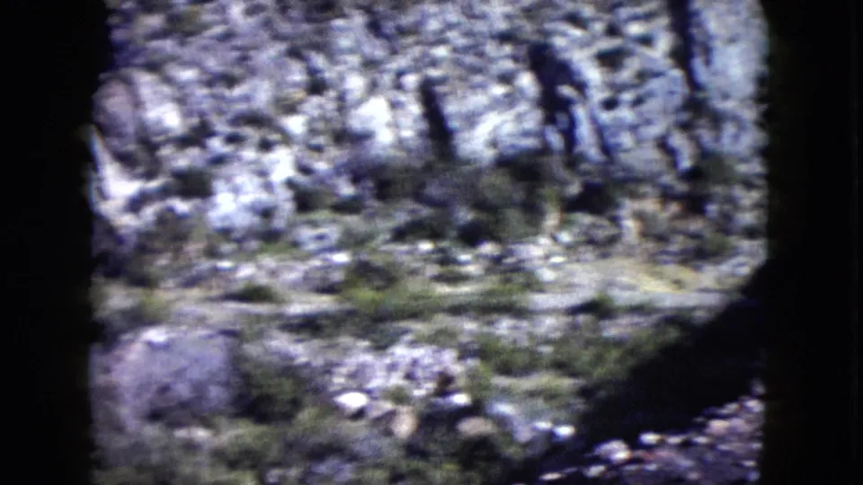 Home-movie view of rocky Nevada desert ground, scattered scrub, and canyon textures within a soft, grainy 8mm frame.
