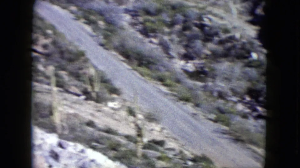 1960 Nevada 8mm frame showing a narrow paved road curving through rocky desert hills with sparse vegetation.
