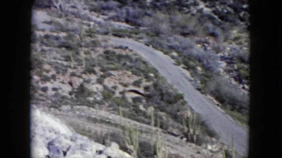1960 Nevada 8mm frame showing a winding road through rocky, dry hills with scattered desert vegetation.