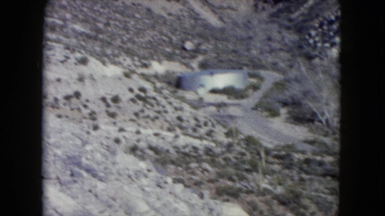 1960 Nevada desert view with rocky ground, sparse shrubs, and a distant light-colored structure or roadside feature.