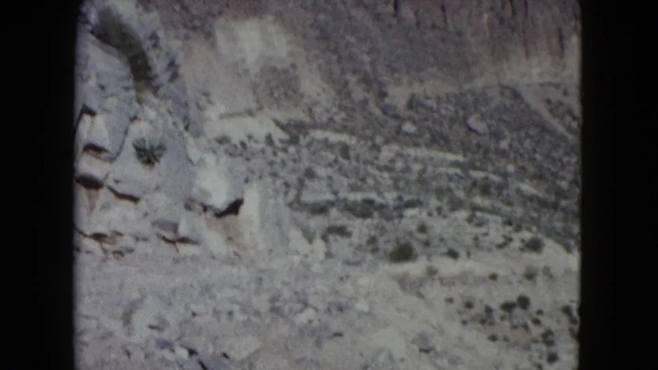 1960 Nevada 8mm frame showing pale rock faces, arid hillside, and scattered desert vegetation.