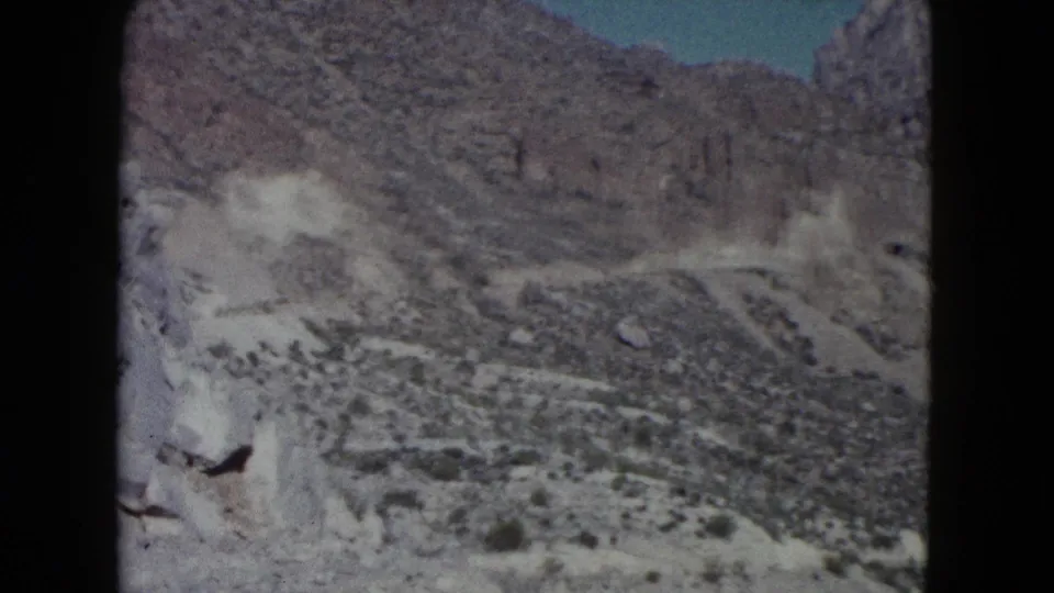 1960 Nevada desert landscape with pale cliffs, a winding road, rocky hills, sparse vegetation, and a small patch of sky.