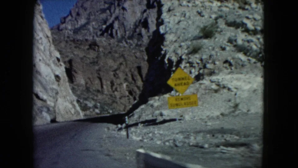 1960 Nevada road-trip frame with a yellow warning sign, paved road edge, gravel shoulder, and steep rocky canyon walls.