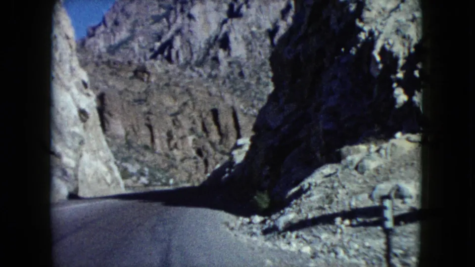 1960 Nevada 8mm view of a curving road entering shadow between steep desert rock walls and rough roadside gravel.