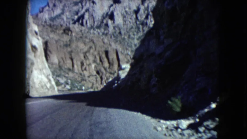 1960 Nevada 8mm road-trip frame showing a paved curve through rocky desert mountains from a moving car perspective.