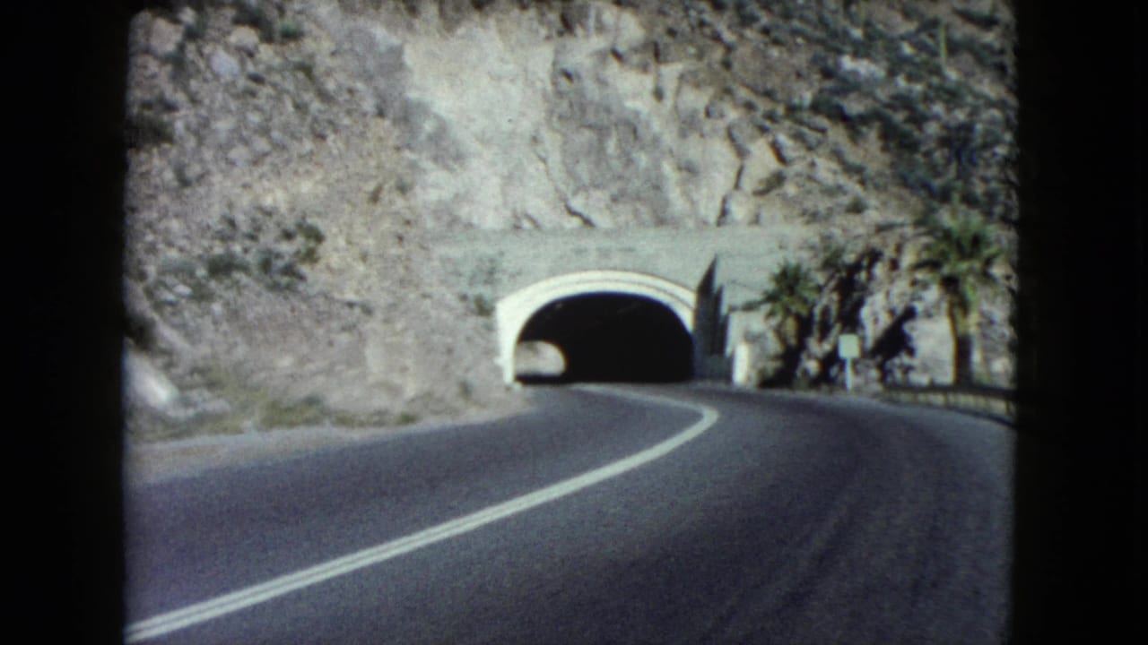 A Nevada mountain road curves toward a pale tunnel portal, with desert rock and palm trees near the entrance.