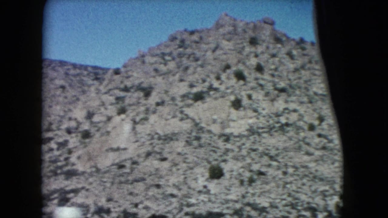 1960 Nevada 8mm frame showing a rocky desert hillside, sparse vegetation, blue sky, and dark window edges from a moving car.
