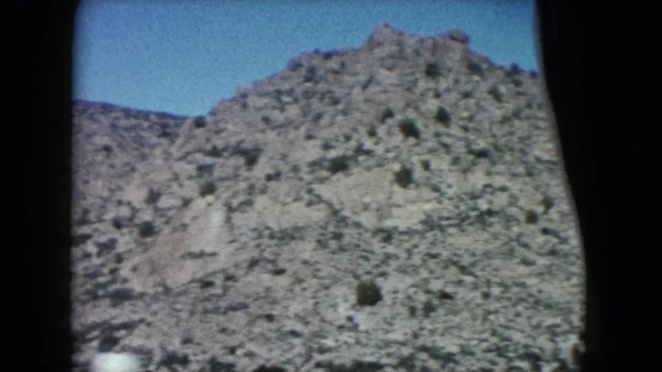 1960 Nevada 8mm frame showing a rocky desert hillside, sparse vegetation, blue sky, and dark window edges from a moving car.