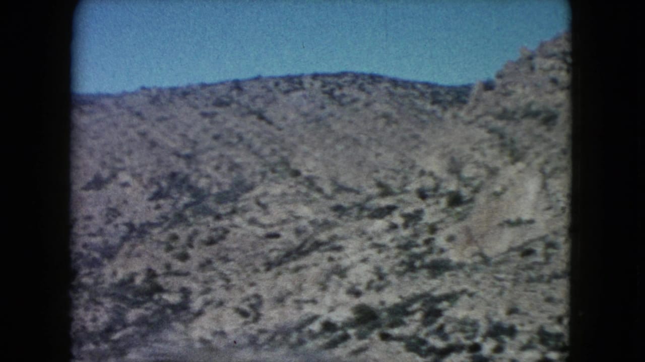 A scrub-covered Nevada ridge passes across the frame, with rocky terrain and soft 8mm color visible through the car-window view.