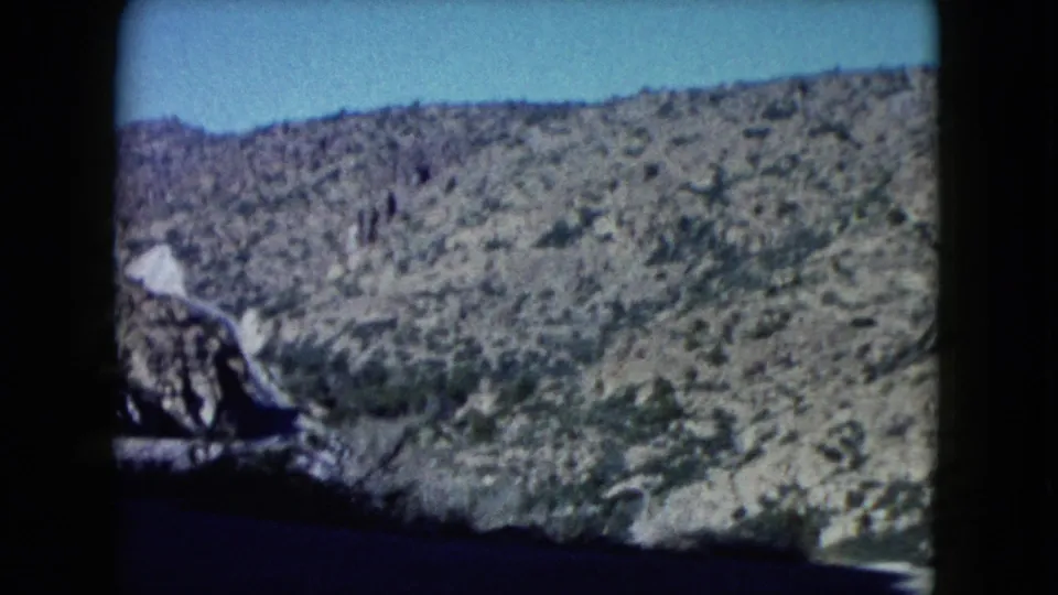 1960 home-movie view of Nevada mountain terrain with scrub vegetation, rocky slopes, blue sky, and shadowed car-window edges.