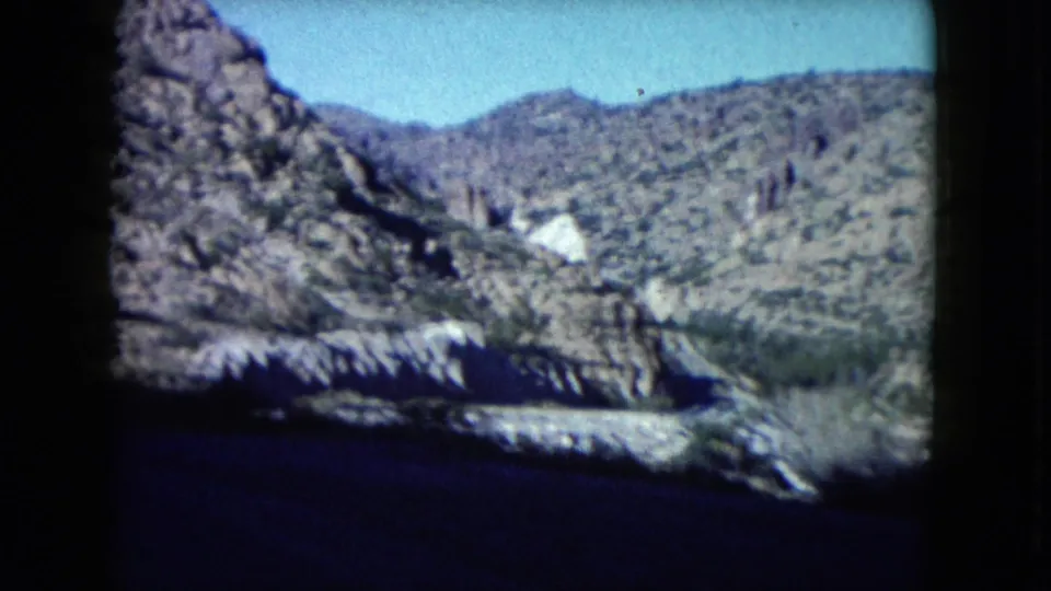 Nevada 8mm travel frame showing rocky hills, sparse desert growth, bright sky, and a dark foreground area from the car-window perspective.