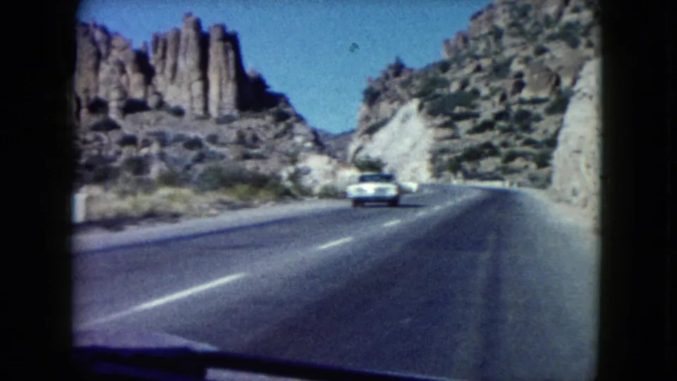 1960 Nevada road-trip footage shows a white car ahead on a winding highway bordered by rugged rock formations.