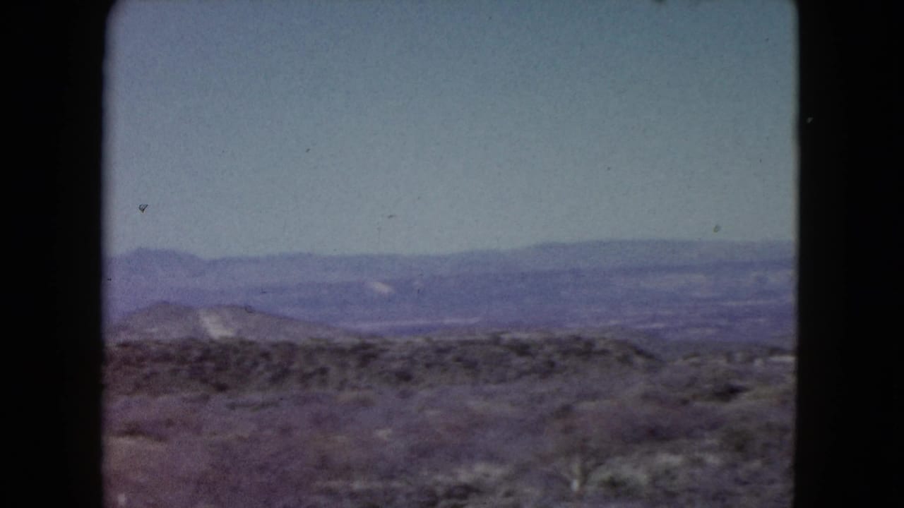 1960 Nevada 8mm frame showing scrubby desert foreground, low hills, and hazy distant mountains beneath a pale blue sky.