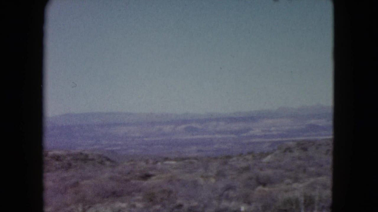 1960 Nevada home-movie view across open desert terrain toward purple-hazed mountain ranges and a clear pale sky.