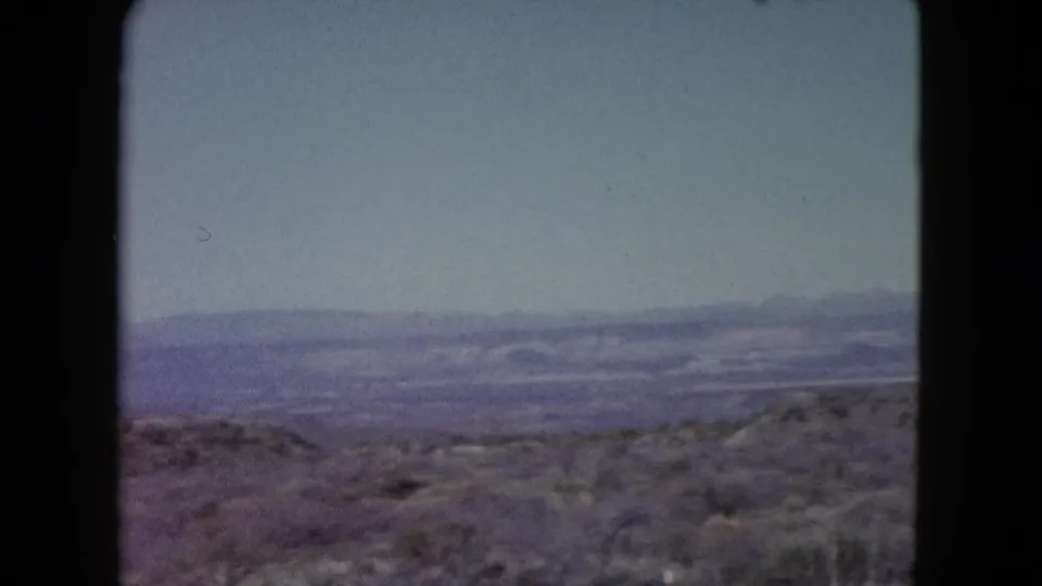 1960 Nevada home-movie view across open desert terrain toward purple-hazed mountain ranges and a clear pale sky.