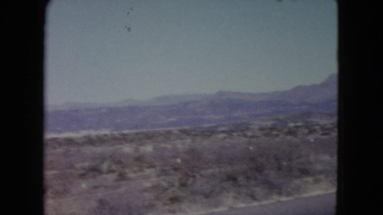 1960 Nevada home-movie frame showing arid roadside scrub and hazy mountains beyond the desert plain.