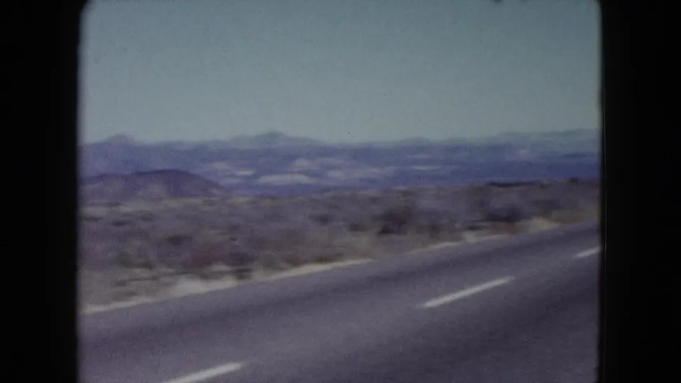 Moving 8mm view along a Nevada highway, with desert shoulder, low brush, and mountains on the horizon.