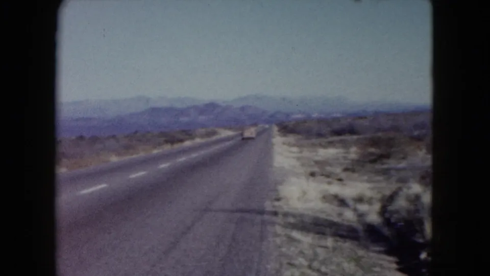 Driver’s-eye 1960 Nevada road-trip view of a highway running through desert scrub toward hazy mountains.