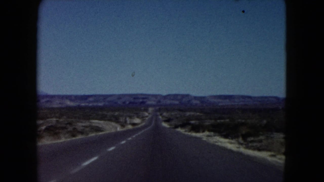 1960 Nevada home-movie frame showing a straight paved desert highway stretching toward low distant hills, with scrubland on both sides and blue sky above.