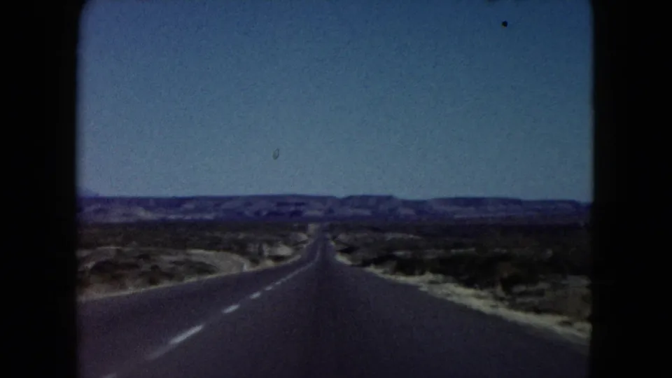 1960 Nevada home-movie frame showing a straight paved desert highway stretching toward low distant hills, with scrubland on both sides and blue sky above.