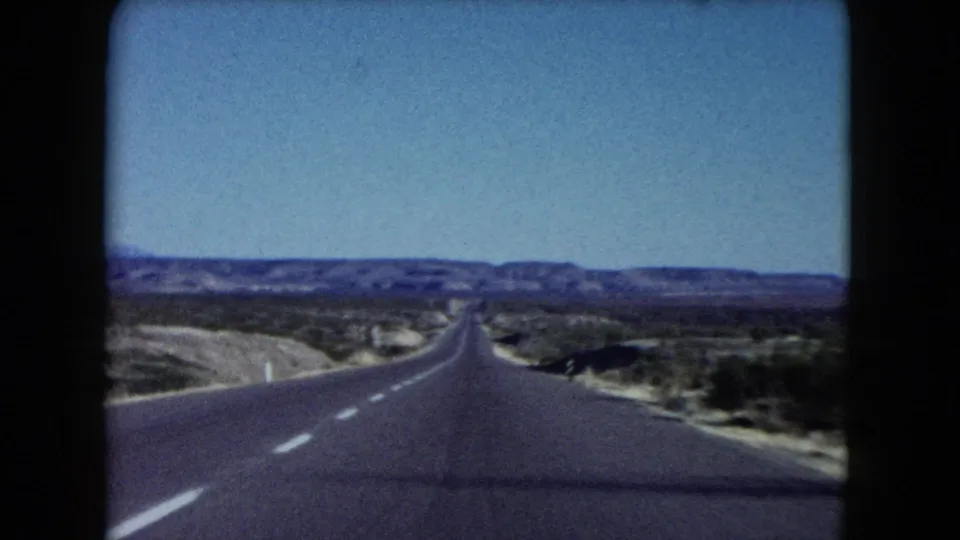 1960 Nevada road-trip footage frame with an empty paved highway, pale shoulders, scattered desert vegetation, and purple-toned mountains on the horizon.