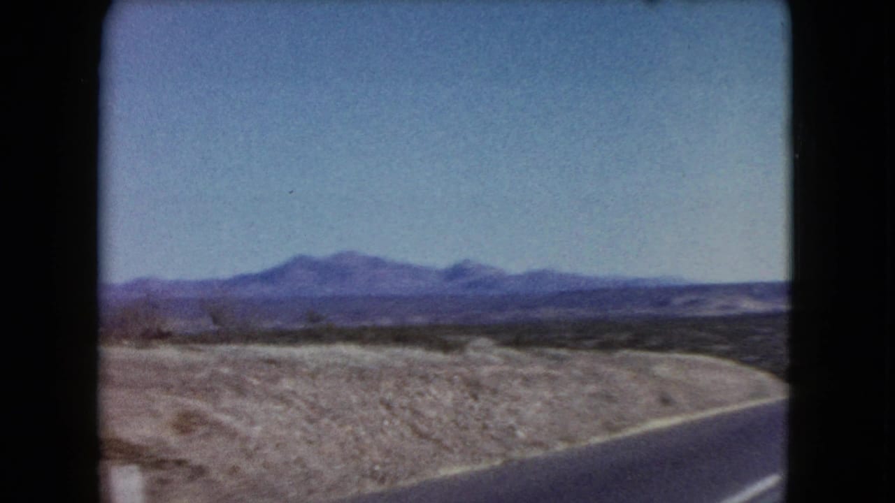 1960 Nevada home-movie frame showing the edge of a paved road curving through open desert, with scrub-covered ground and low mountains beyond.