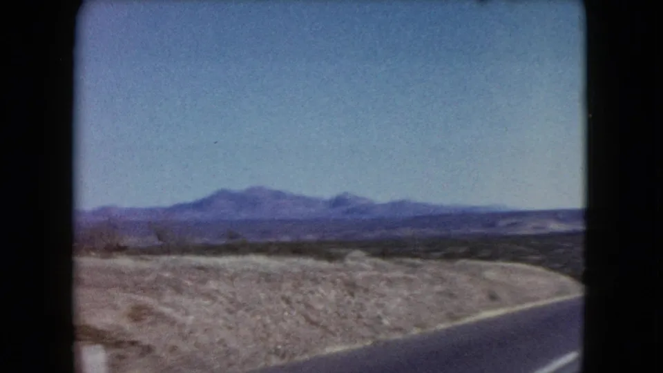 1960 Nevada home-movie frame showing the edge of a paved road curving through open desert, with scrub-covered ground and low mountains beyond.