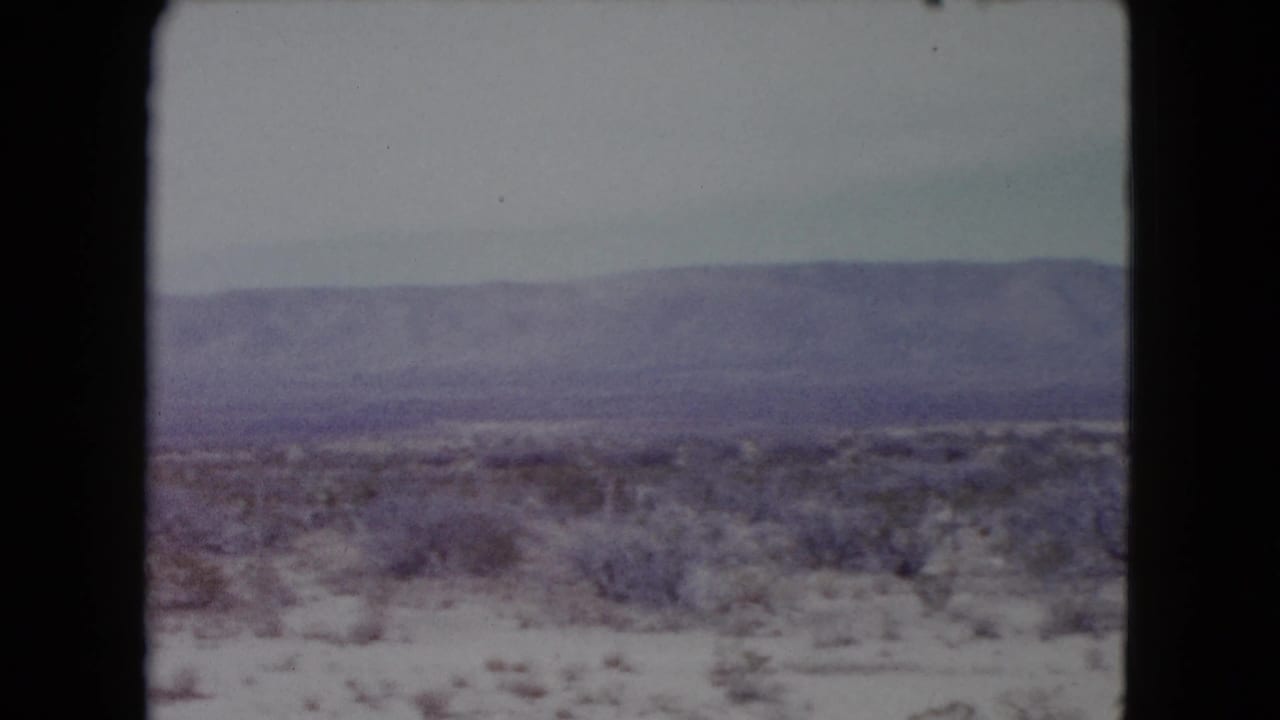 1960 Nevada desert travel frame showing scrubland beside a pale roadside area or track, with a broad hazy mountain range in the background.