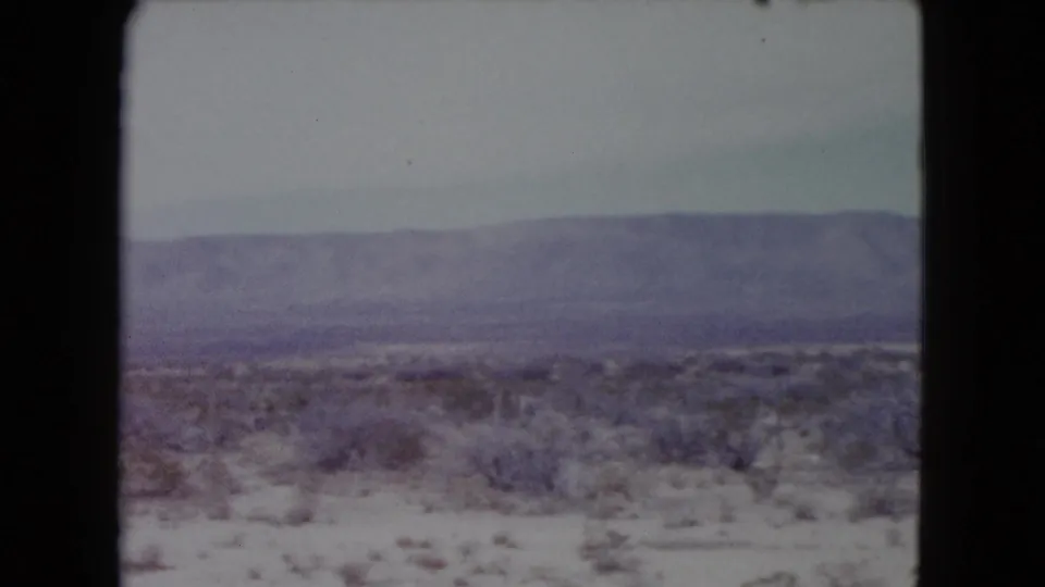 1960 Nevada desert travel frame showing scrubland beside a pale roadside area or track, with a broad hazy mountain range in the background.