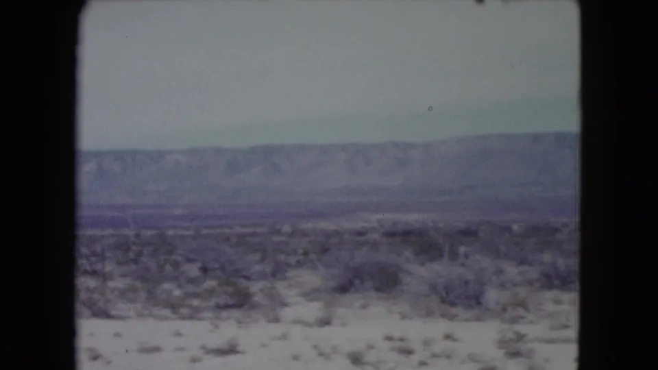1960 Nevada 8mm frame showing a dry scrub-covered plain with a hazy mountain range in the distance.